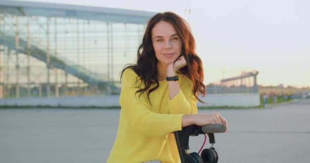 Portret van goed uitziende positieve zorgeloze stijlvolle jonge brunette in gele trui die in de buurt van een eigen e-scooter op de achtergrond van de moderne panoramische luchthaven overdag — Stockvideo