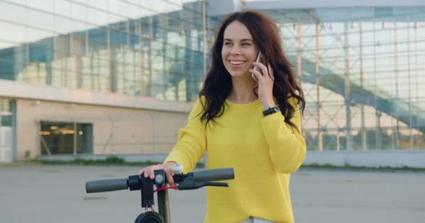 Bella sorridente felice giovane donna alla moda in maglione giallo a piedi con e-scooter e parlare su smartphone vicino a grande edificio moderno durante il giorno — Video Stock