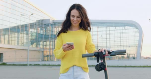 Retrato de una mujer de pelo largo alegre positiva y elegante que usa su teléfono mientras camina con e-scooter cerca de un gran edificio urbano en verano — Vídeo de stock