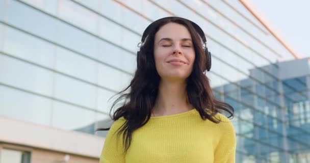 Splendida in spiriti alti sorridente giovane donna elegante in maglione giallo godendo melodie preferite in cuffie vicino edificio di vetro panoramico nella giornata di sole, rallentatore — Video Stock