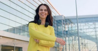 Gündüz vakti panoramik cam binanın yanında çapraz kollarla poz veren güzel, güler yüzlü, zarif, şık, genç bir esmerin portresi.