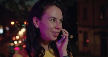 Cazibeli, güler yüzlü, uzun saçlı, neşeli genç bir bayana yakın ol. Gecenin ışıklarının arasında telefonla konuşuyor.