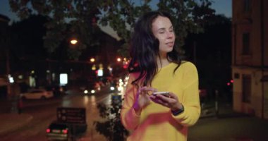 Güzel görünüşlü, kaygısız, koyu renk saçlı, sarı kazaklı bir kadın akıllı telefonunu şehrin renkli ışıkları arasında kullanıyor.