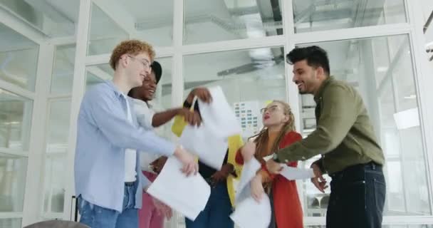 Good-looking friendly smiling experienced young diverse group of office workers rejoicing successful ending joint project and throwing up documents over their heads in modern meeting room — Stock Video