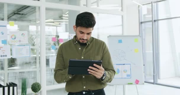 Good-looking confident purposeful high-skilled hispanic guy standing in modern office room and working on tablet pc,front view — Stock Video