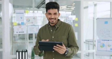 İyi huylu, başarılı, yaratıcı, yetenekli İspanyol çocuk elinde tablet bilgisayar tutarken modern cam ofis odasında poz veriyor.