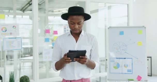 Retrato de atractivo concentrado joven negro exitoso en ropa elegante y sombrero que trabaja en la computadora de tableta en la sala de reuniones moderna — Vídeos de Stock