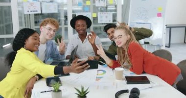 Güzel görünüşlü, kendine güvenen, stil sahibi, çok ırklı arkadaşlar ofis masasında oturuyor ve ortak çalışma sırasında cep telefonunda grup fotoğrafı çekiyor. Kapat.