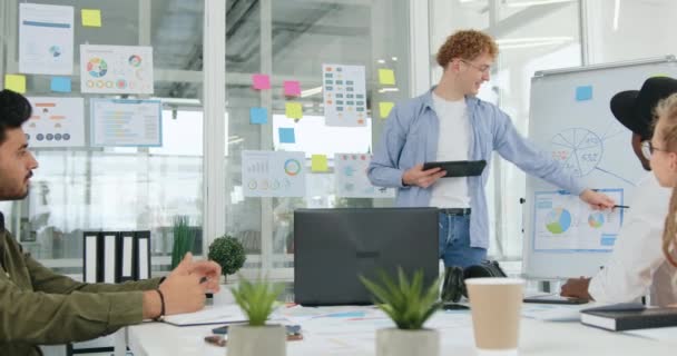 Simpático sonriente confiado hombre hábil con el pelo rizado celebración de una presentación para sus colegas profesionales de raza mixta con experiencia y el análisis con ellos infografías — Vídeos de Stock