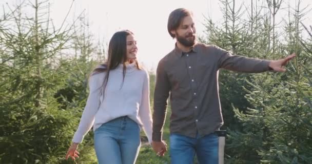 Närbild av godmodiga positiva unga leende man och kvinna som vandrar bland planteringar med x-mas träd i skogsbruk när de kommer att välja traditionella träd för semester — Stockvideo