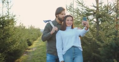 Çekici, mutlu, tutkulu, genç bir çift köknar ağaçlıklarından birini tatil için kesmeye geldiklerinde telefonda resim çekiyorlar.