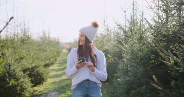 Çekici, mutlu, güler yüzlü, sıradan elbiseli ve şapkalı genç bir kadın köknar ağaçlarının arkasında duruyor ve akıllı telefonunu kullanıyor. — Stok video