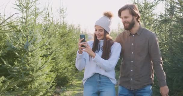 気分が良いです現代的な30代のカップルで愛歩くの行の間にモミの木の植え付けとリビジョン写真で彼女の携帯電話,閉じる — ストック動画