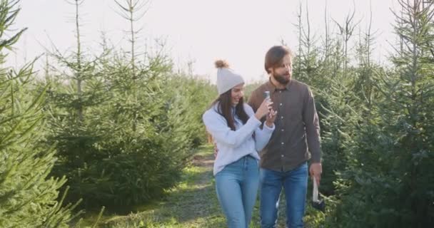 Attraente positivo felice giovane uomo barbuto e sua moglie è venuto in un posto speciale con gli impianti di abeti per tagliare l'albero per loro, facendo foto per scegliere la migliore bellezza della foresta per le vacanze — Video Stock