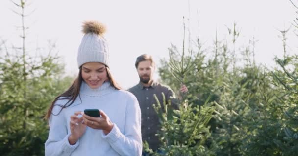 Härligt leende ung kvinna i hatt med hjälp av sin telefon när nöjd skäggig make förde vacker gran träd som han väljer på särskild plats i skogen — Stockvideo