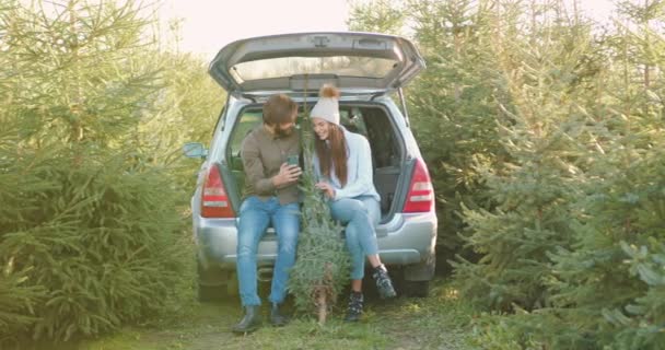 Приємна щаслива молода пара сидить у багажнику автомобілів серед зелених ялин та переглядає фотографії на смартфоні, крупним планом, повільний рух — стокове відео