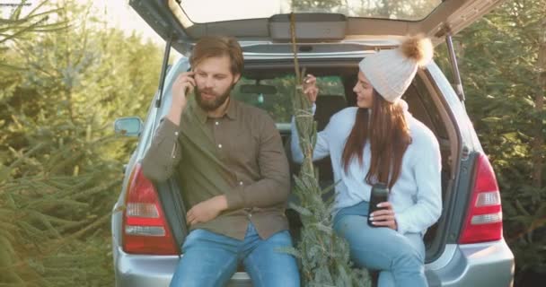 Snygg glad leende ung kvinna sitter tillsammans med sin glada skäggig pojkvän i bilar bagagelucka som talar på smartphone efter att hur de köpte gran i skogen — Stockvideo
