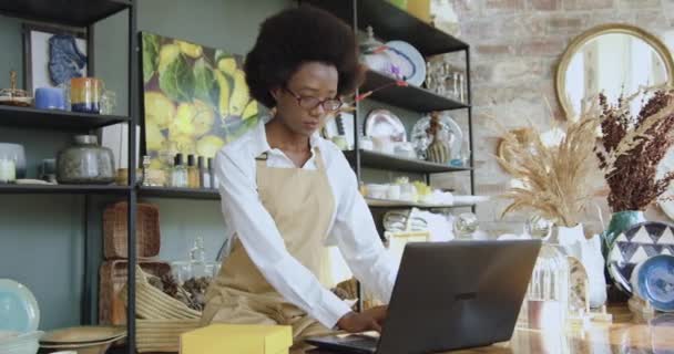 Atractiva mujer concentrada segura de sí misma de piel negra con peinado africano escribiendo en el portátil durante el trabajo en la tienda de recuerdos — Vídeos de Stock