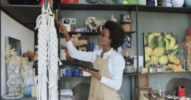 Oldukça çalışkan, sakin, siyah tenli bir kadın hediyelik eşya dükkanında mal satıyor ve verileri tablet bilgisayara, yavaş çekimde yazıyor.