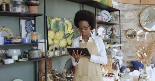 매력적 인 미소를 짓고 있는 젊은 아프리카계 미국인 여성이 손으로 만든 아름다운 기념품 가게에서 태블릿을 만들고 있고 행운의 얼굴로 카메라를 보고 있습니다. — 비디오