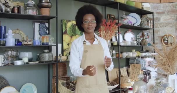 Encantadora mujer afroamericana feliz sonriente en blusa blanca con delantal posa en la cámara con los brazos cruzados en su tienda de regalos hecha a mano, cámara lenta — Vídeos de Stock