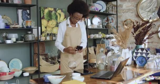 Вид спереди на довольно уверенную трудолюбивую молодую темнокожую женщину в фартуке, которая работает на ноутбуке в красивом сувенирном магазине ручной работы — стоковое видео