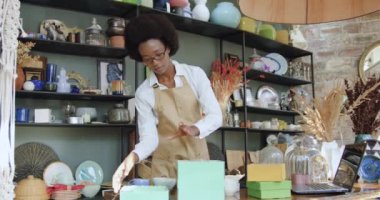 Güzel bir hediyelik eşya dükkanında çalışan ve şarap kadehlerini ve bardakları hediye kutularına dolduran kendine güvenen Afro-Amerikan kadının ön görüntüsü.