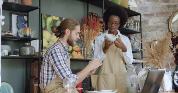 Aangename positieve hardwerkende gemengde ras mannelijke en vrouwelijke verkopers in cadeauwinkel controleren op slaan of kraken kopjes en het invoeren van hun beschrijvingen in computerrapport — Stockvideo