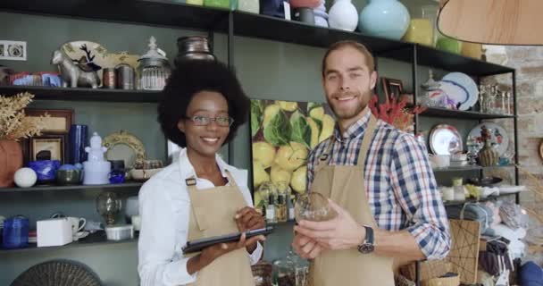 Portrett av attraktive unge, smilende raseselgere som står foran kamera i koselig gavebutikk og viser tomler opp – stockvideo