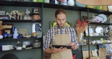Önlüklü 30 yaşında gülümseyen güzel görünümlü sakallı satıcı tablet cihazı kullanıyor ve harika bir hediyelik eşya dükkanında çalışırken kameraya bakıyor. Kapatın.