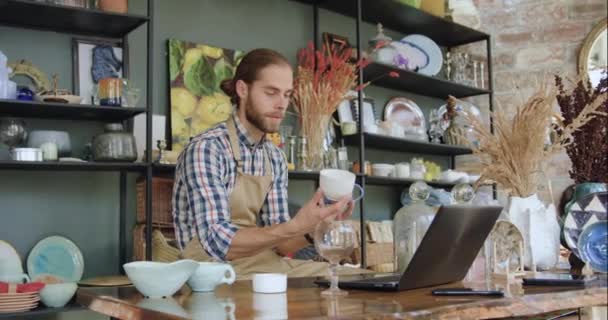 Вероятный босоногий продавец в фартуке, сидящий на рабочем месте и сравнивающий голубую чашку с белой во время работы в красивом сувенирном магазине — стоковое видео