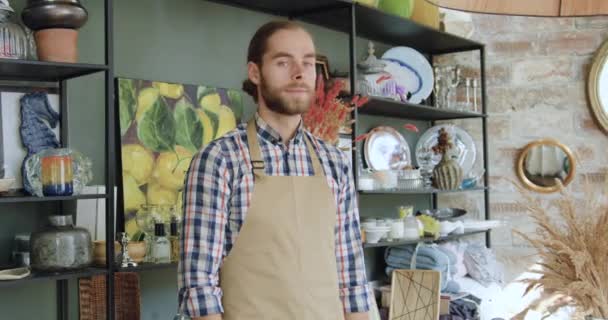 Aangename tevreden jonge bebaarde winkelier in schort kijken naar camera met gekruiste armen tijdens het werken in prachtige kleine cadeauwinkel, close-up — Stockvideo