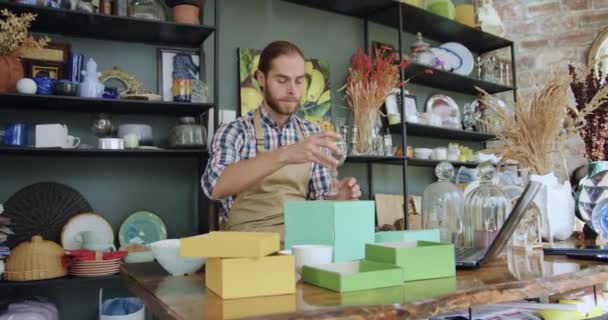 Front view of likable hardworking young bearded guy which packing wine glass into gift box while working in beautiful gift shop — Stock Video