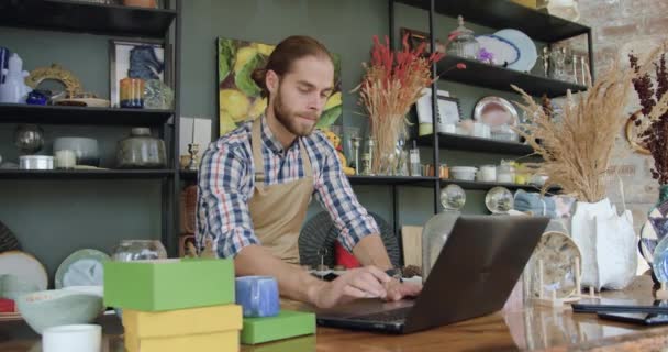 Доброзичливий задоволений усміхнений молодий бородатий чоловік в фартусі сидить перед комп'ютером в красивому сувенірному магазині і дивиться на екран з щасливим обличчям — стокове відео