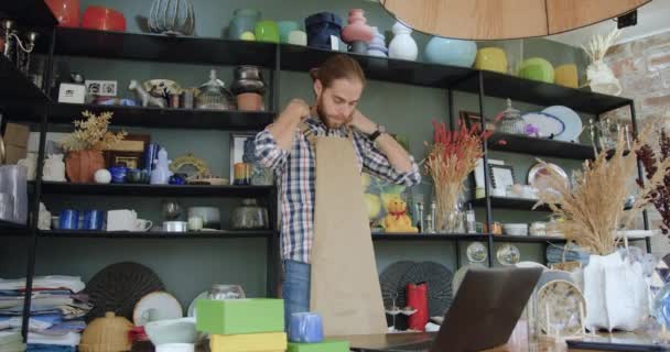 Close up of good-looking confident modern young bearded man which ties his work apron before starting to work in contemporary handmade gift shop — Stock Video