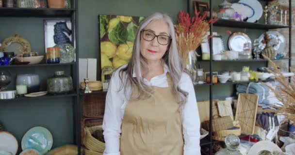 Retrato de adorable sonriente mujer madura feliz con pelo gris en gafas que posa en cámara con brazos cruzados en hermosa tienda de regalos — Vídeos de Stock