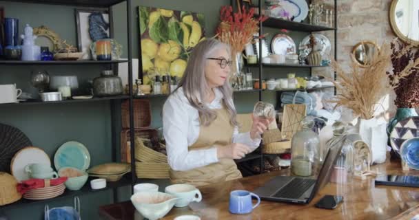 Attractive confident grey haired mature woman in glasses sitting at the computer and comparing articles from cozy gift shop and availability on online shopping — Stock Video