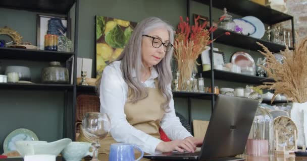 Хорошо выглядящая спокойная седовласая женщина в очках, работающая на ноутбуке в хорошо оборудованном современном сувенирном магазине, замедленная съемка — стоковое видео