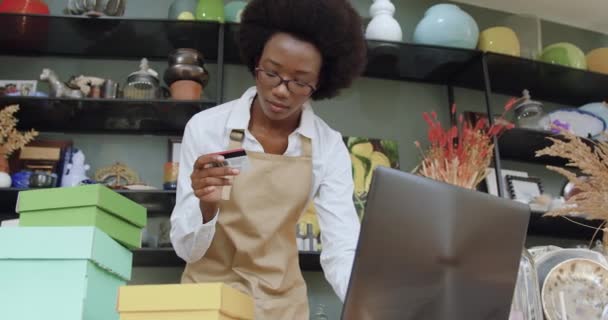 Крупним планом добре виглядає сучасна молода чорношкіра жінка, яка працює в чудовому сувенірному магазині і вводить дані з банківської картки в ноутбук для клієнтів для онлайн-шопінгу — стокове відео