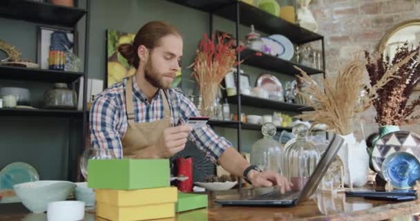 Соответствующий сконцентрированный современный бородатый молодой работник из сувенирного магазина, вводящий данные банковских карт на ноутбуке для покупателей интернет-магазинов, сидящих на своем рабочем месте — стоковое видео