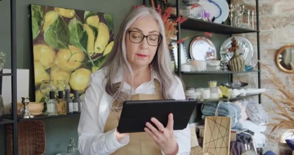 Widok z przodu bardzo uśmiechnięta szczęśliwa starsza pani w okularach, która pozuje przed kamerą z tabletem pracując nad nim w przytulnym sklepie z pamiątkami — Wideo stockowe