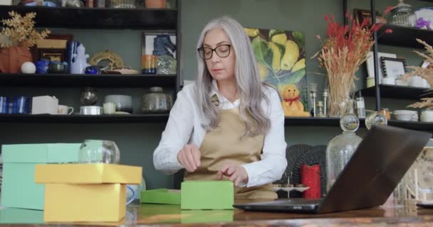 Portrait d'une belle femme âgée confiante et travailleuse dans des lunettes qui met une tasse blanche dans une boîte-cadeau, assise à la table dans une boutique de cadeaux — Video