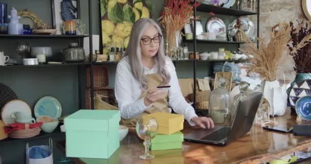 Assez calme prudent vieille femme aux cheveux gris vendeur assis à la table dans la belle boutique de souvenirs et entrer les détails de la carte bancaire dans l'ordinateur pour les clients en ligne — Video