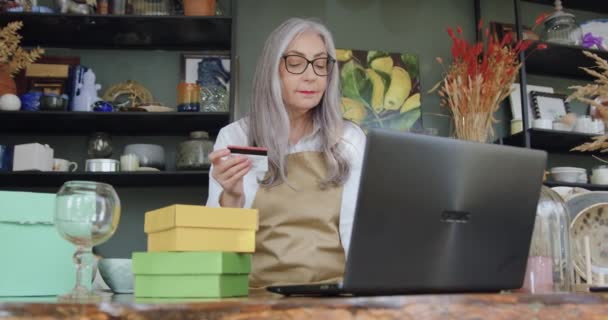 Atractiva de buen humor mujer madura madura de pelo gris sentada frente a la computadora e introduciendo detalles de la tarjeta bancaria para potenciales compradores en línea de bienes de su tienda de regalos — Vídeos de Stock