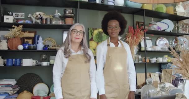 Vista frontal de las mujeres de raza mixta de buen aspecto en altos espíritus sonriendo diferentes edades de la tienda de regalos que posan en la cámara con los brazos cruzados — Vídeos de Stock