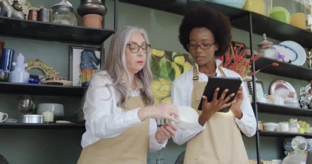 Attraktivt positiv positiv äldre kvinnlig ägare till presentbutik förklara för sin unga afrikanska amercian medarbetare detaljer om gemensamt arbete, slow motion — Stockvideo