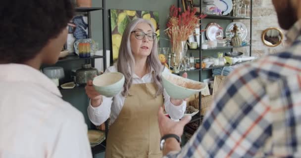 Dobře vypadající přátelský šedovlasý zralý prodejce navrhující krásné nádobí mladému smíšenému páru, který přišel v obchodě se suvenýry, pohled zepředu — Stock video
