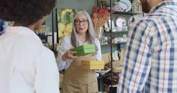 Okouzlující usměvavá zralá žena s šedými vlasy v brýlích dává dárkové krabice sympatické mladé přátelské smíšené rasy turistů, které si koupili v obchodě se suvenýry — Stock video