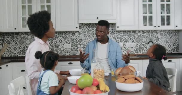 Concepto familiar donde adorables padres felices sonrientes y sus niñas pequeñas y agradables sentados alrededor de la mesa de la cena en la cocina contemporánea y hablando durante el desayuno, cámara lenta — Vídeos de Stock