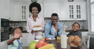 Çekici, gülümseyen, siyah tenli, mutlu bir ailenin ön görüntüsü. Çağdaş mutfakta kahvaltıda tatmin olmuş yüzleriyle kameraya bakan iki güzel kız.